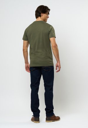 Tricou scurt cu mânecă în verde măsliniu, din bumbac, cu o croială lejeră, asortat cu blugi de culoare albastru închis și încălțăminte maro, vazut din spate.