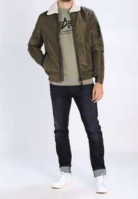 Giacca bomber verde oliva con colletto in shearling bianco, abbinata a una t-shirt grigia, jeans scuri e sneakers bianche.