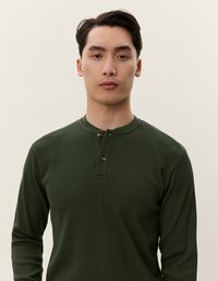 Hosszú ujjú, bordázott henley póló sötétzöld színben, lekerekített nyakkivágással és két gombos zipzárral, texturált anyagból készült.