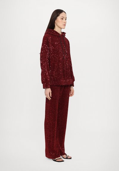 Red sequined hoodie and matching pants, featuring a drawstring hood, relaxed fit, and textured surface. Worn with black sandals.
