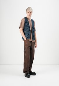 Joven con pelo corto rubio que está de pie, vestido con una camiseta marrón, chaleco de mezclilla con bolsillos, pantalones marrones holgados y zapatos negros gruesos.