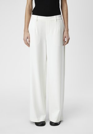 Vrouw draagt witte, brede broek met hoge taille en voorzakken, gecombineerd met zwarte schoenen, staand tegen een effen achtergrond.