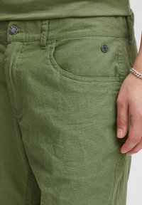 Žalios spalvos lino kelnės su tekstūruota viršutine dalimi, su priekinėmis kišenėmis ir sagų užsegimu. Priartintas vaizdas atskleidžia audinio minkštumą ir spalvą.