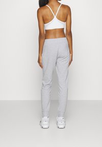 Bílá sportovní podprsenka s designem na zádech ve tvaru racerback v kombinaci se šedými tepláky. Oblečení má měkkou texturu a pohodlný střih, doplněné bílými teniskami.