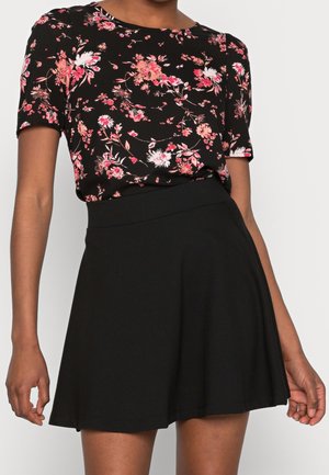 Frau trägt ein schwarzes kurzärmeliges Oberteil mit pink-weißem Blumenmuster, kombiniert mit einem einfarbigen schwarzen Rock in A-Linie.