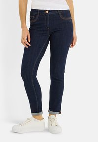 Jean en denim bleu foncé à coupe slim, avec coutures visibles et revers roulés. Associé à des baskets blanches et un haut de couleur claire.