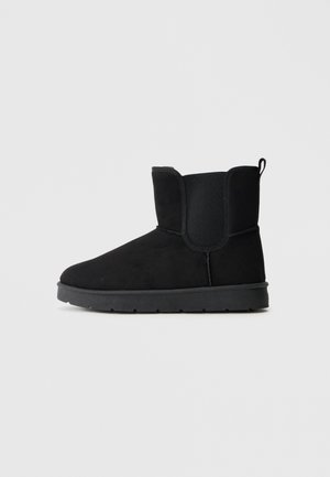 Botte d’hiver noire à hauteur de cheville avec semelle plate, embout arrondi, panneaux latéraux élastiques et languette au talon, présentée de profil sur fond uni.