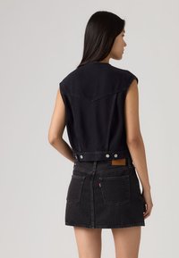 Γυναίκα που φοράει αμάνικο μαύρο τζιν μπουφάν και μαύρη τζιν φούστα, με θέα από πίσω, πάνω σε απλό φωτεινό φόντο.