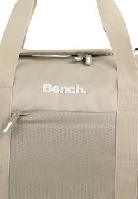 Beige weekendtas van gladde stof, met een hexagonaal patroon op de voorzak, bovenaan afsluitbaar met een rits en een wit merklogo. Duurzame webbing handgrepen.