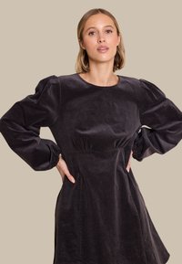 Mørk kordfløyelskjole med puffer lange ermer, rund hals og innsvingt midje. Glatt tekstur med subtile vertikale ribber.