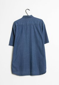 Camicia in denim a maniche corte, colletto a bottoni, cucitura del yoke posteriore e orlo curvo. Colore blu scuro con una texture liscia. Appesa a un gancio nero.