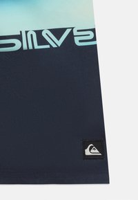 Υφάσματα σε ναυτικό και ανοιχτό μπλε χρώμα με μερικό λογότυπο "Quiksilver" και μικρό ραμμένο μαύρο σημάδι που απεικονίζει ένα λευκό έμβλημα με κύμα και βουνό.