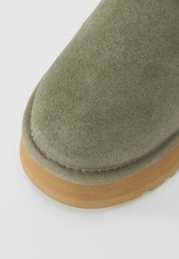 Close-up of a green suede shoe toe with beige rubber sole and fabric trim on a white background.