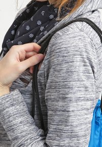 Osoba v šedé mikině s kapucí a námořnickém šálu s námořními vzory, která drží černý popruh přehozený přes rameno.