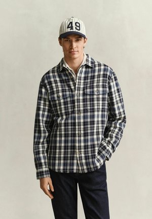 Jeune homme portant une chemise à carreaux blanche et bleu marine, un jean foncé et une casquette blanche avec le numéro 49, debout avec une main dans la poche.
