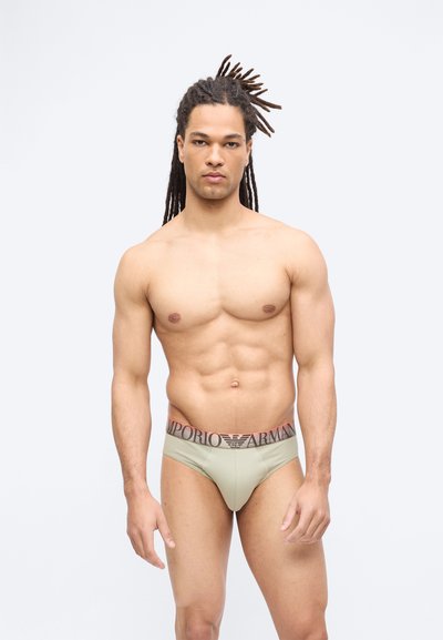 Μυώδης άνδρας με μακριές κοτσίδες φοράει μπεζ εσώρουχα Emporio Armani, στέκεται μπροστά σε απλό φωτεινό φόντο με ουδέτερη έκφραση.