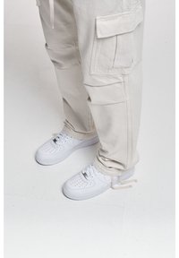 Jambes portant un pantalon cargo beige et des baskets blanches, debout sur un sol blanc, les lacets de la chaussure droite dénoués.