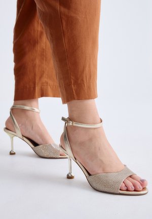 Pies femeninos con tacones altos dorados brillantes y abiertos en la punta con correas en el tobillo, combinados con pantalones cortos de color óxido.