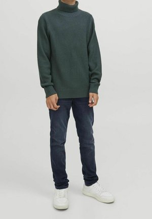 Personne portant un pull à col roulé en tricot gaufré vert foncé, un jean bleu foncé et des baskets blanches, debout devant un fond uni.