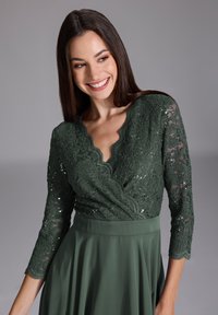 Robe en dentelle verte à manches trois-quarts, décolleté festonné et corsage ajusté, se prolongeant en une jupe plissée. Dentelle texturée et éclat subtil.