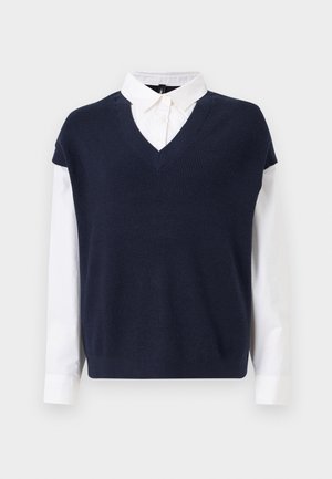 Pull en V navy superposé à une chemise blanche à col. Le pull présente une texture côtelée ; la chemise a des manches longues et des poignets à boutons.