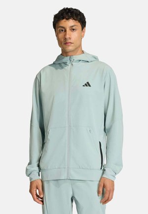 Uomo che indossa una giacca con cappuccio e zip azzurro chiaro e pantaloni abbinati con logo nero sul petto, su sfondo uniforme.