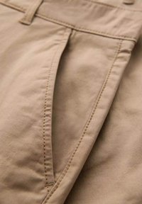 Close-up af lomme på beige bukser med synlige syninger og del af linning med knap.