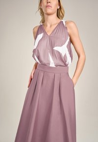 Ärmelloses Kleid mit V-Ausschnitt, das ein gefaltetes, helllila Oberteil und einen festen, weiten Rock mit Taschen zeigt, ergänzt durch weiße abstrakte Muster.