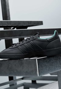 Baskets basses noires avec trois bandes et doublure intérieure bleu sarcelle, reposant sur un banc en métal avec un arrière-plan industriel flou.