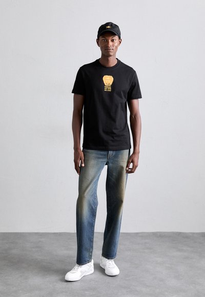 Schwarzes T-Shirt mit goldenem Löwen-Graphic, blaue Jeans mit leichter Ausbleichung und weiße Sneakers. Schwarze Mütze mit einem Logo. Der Pose des Models wird eingenommen.