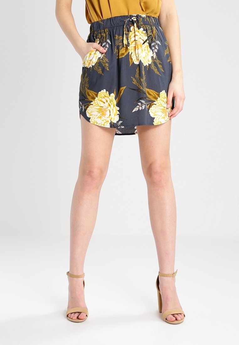 Femme portant une mini-jupe bleue marine à fleurs jaunes, un haut moutarde et des sandales à talons hauts beiges, debout avec une main dans la poche.