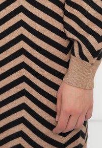 Nærbillede af en hånd, der hviler på et strikket plagg i beige og sort med zigzag mønster og ribkant.