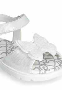 White sandal featuring a shiny metallic strap, textured fabric, and a large white butterfly decoration on a heart-patterned sole.