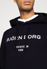 Felpa con cappuccio nera con testo stampato bianco "BJÖRN BORG STOCKHOLM 1956." Presenta lacci e un tessuto morbido e testurizzato.