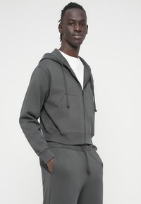 Γκρι φούτερ με φερμουάρ, κουκούλα με κορδόνια και μπροστινές τσέπες, κατασκευασμένο από μαλακό ύφασμα. Συνδυάζεται με γκρι φόρμα που ταιριάζει.