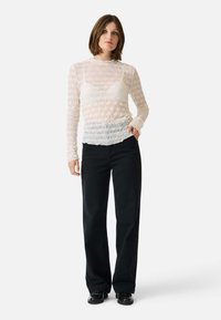 Transparente, texturierte Langarmbluse in hellem Weiß mit gerafftem Muster, kombiniert mit lockeren schwarzen Hosen und schwarzen Schuhen.