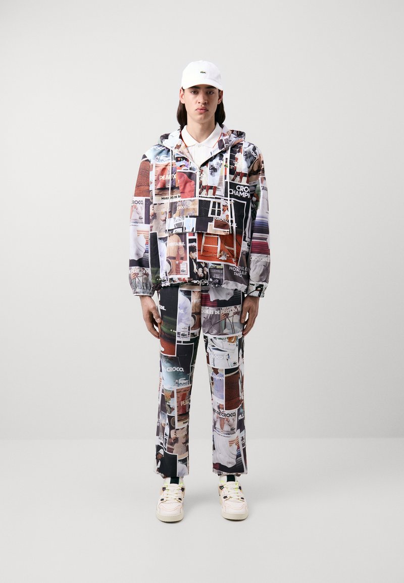 Chaqueta y pantalones impresos que presentan un collage de imágenes coloridas. La tela es ligera y con un acabado brillante. Se lleva con una gorra blanca.