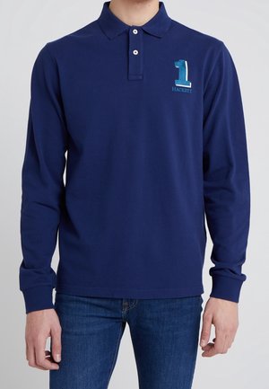 Homme portant un polo bleu marine à manches longues avec le numéro 1 et le logo "Hackett" sur la poitrine, associé à un jean bleu.