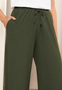 Zelené široké kalhoty s žebrovanou strukturou, které mají elastický pas se stahovací šňůrkou a boční kapsy, vykazující texture látky.
