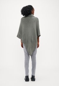 Grå strikket poncho med høy krage, med en trekantet form og vide ermer, som henger ned til midten av ryggen over lysegrå jeans og svarte støvler.