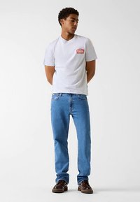 Weißes T-Shirt mit kurzen Ärmeln und rotem "Guess"-Logo, kombiniert mit blauen Jeans und braunen Schuhen. Schlichtes Design, reguläre Passform, Baumwollmaterial.