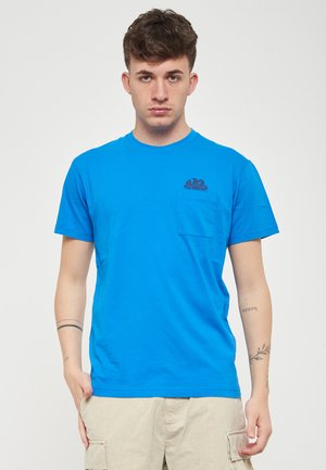 T-shirt in cotone blu ciano con maniche corte e tasca sul lato sinistro del petto, caratterizzato da una grafica di onde blu scuro sopra la tasca.