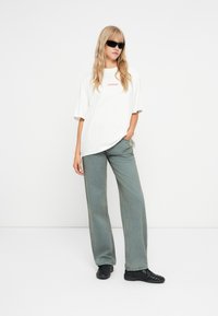 Λευκό oversized t-shirt με την ένδειξη "PEGADOR" σε κόκκινο, συνδυασμένο με πράσινο φαρδύ τζιν και μαύρα αθλητικά παπούτσια. Απαλή ύφανση και άνετη εφαρμογή.