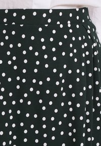 Jupe en tissu vert foncé avec des pois blancs, dotée d'un design plissé et d'une texture lisse. Taille élastique pour un ajustement confortable.