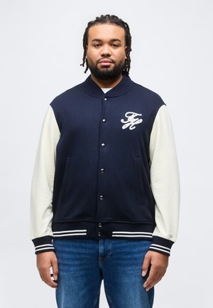 Mann mit geflochtenem Haar, der eine marineblaue Varsity-Jacke mit weißen Ärmeln und dem Buchstaben "FH" auf der Brust sowie eine blaue Jeans trägt, steht vor einem einfarbigen Hintergrund.