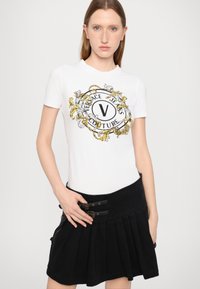 Valge t-särk mustade ja kuldsete detailidega, millel on "Versace Jeans Couture" logo; sobitatud musta kortsunud seelikuga, millel on metallilised aksessuaarid.