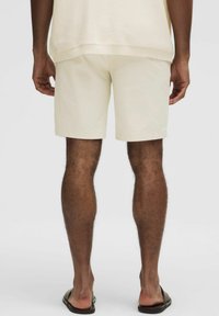 Krémové kraťasy s rovný strihom a hladkou textúrou, skombinované s čiernymi sandálmi. Záber sa zameriava na zadnú časť osoby.