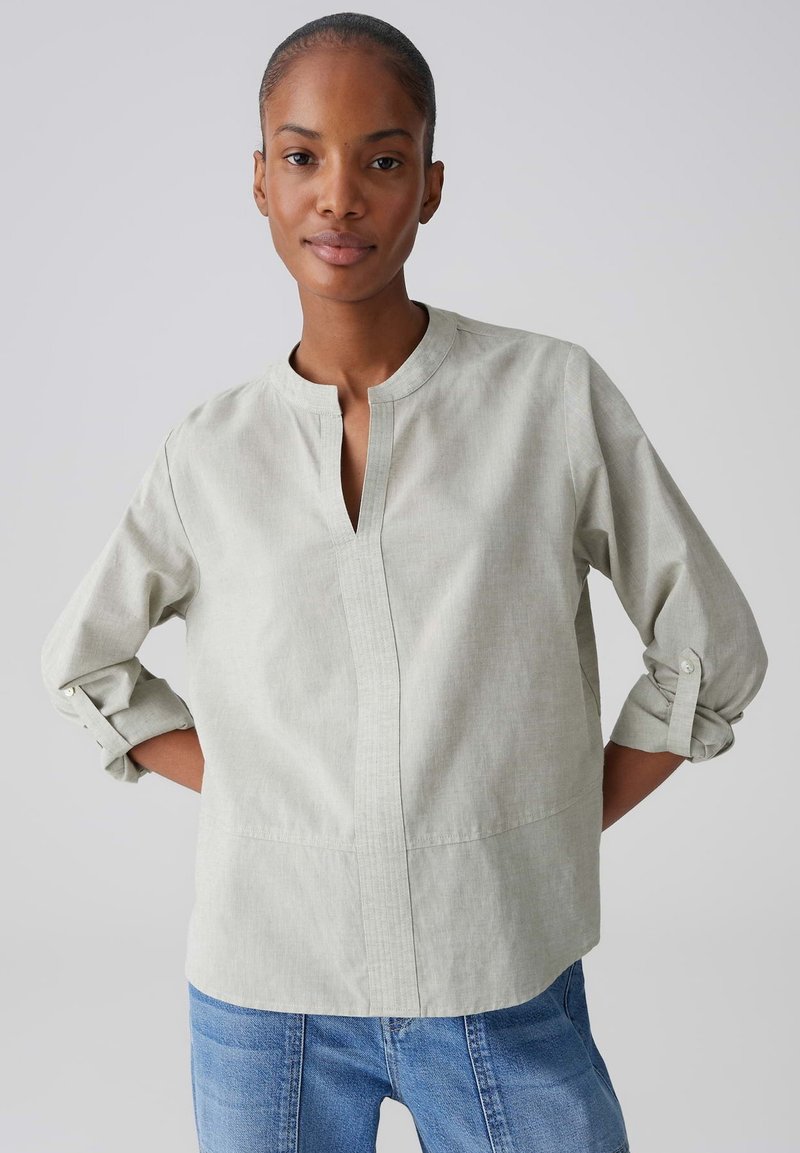 Femme aux cheveux tirés en arrière, portant un chemisier gris clair à manches longues avec les poignets retroussés et un jean bleu, debout devant un fond uni.