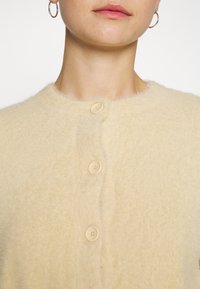 Femme portant un cardigan duveteux de couleur crème, boutonné en haut, avec des boucles d'oreilles en anneau doré visibles aux oreilles.