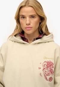 Beżowa bluza z kapturem z czerwonym haftem smoka i tekstem "Superdry". Nałożona na koszulę w kratę z widocznym kołnierzykiem.